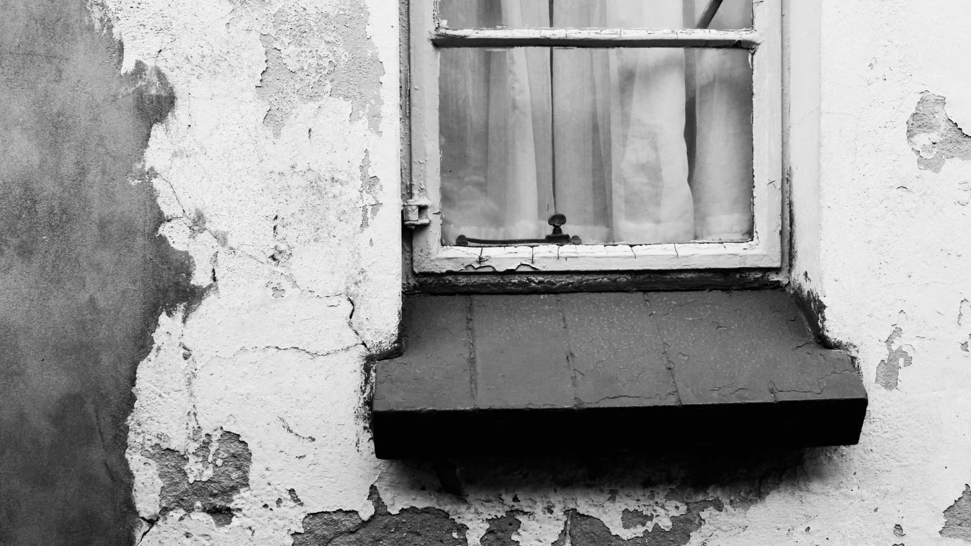 A black-and-white photo of an old building with a white wooden window frame covered by a curtain, and peeling, cracked plaster on the wall